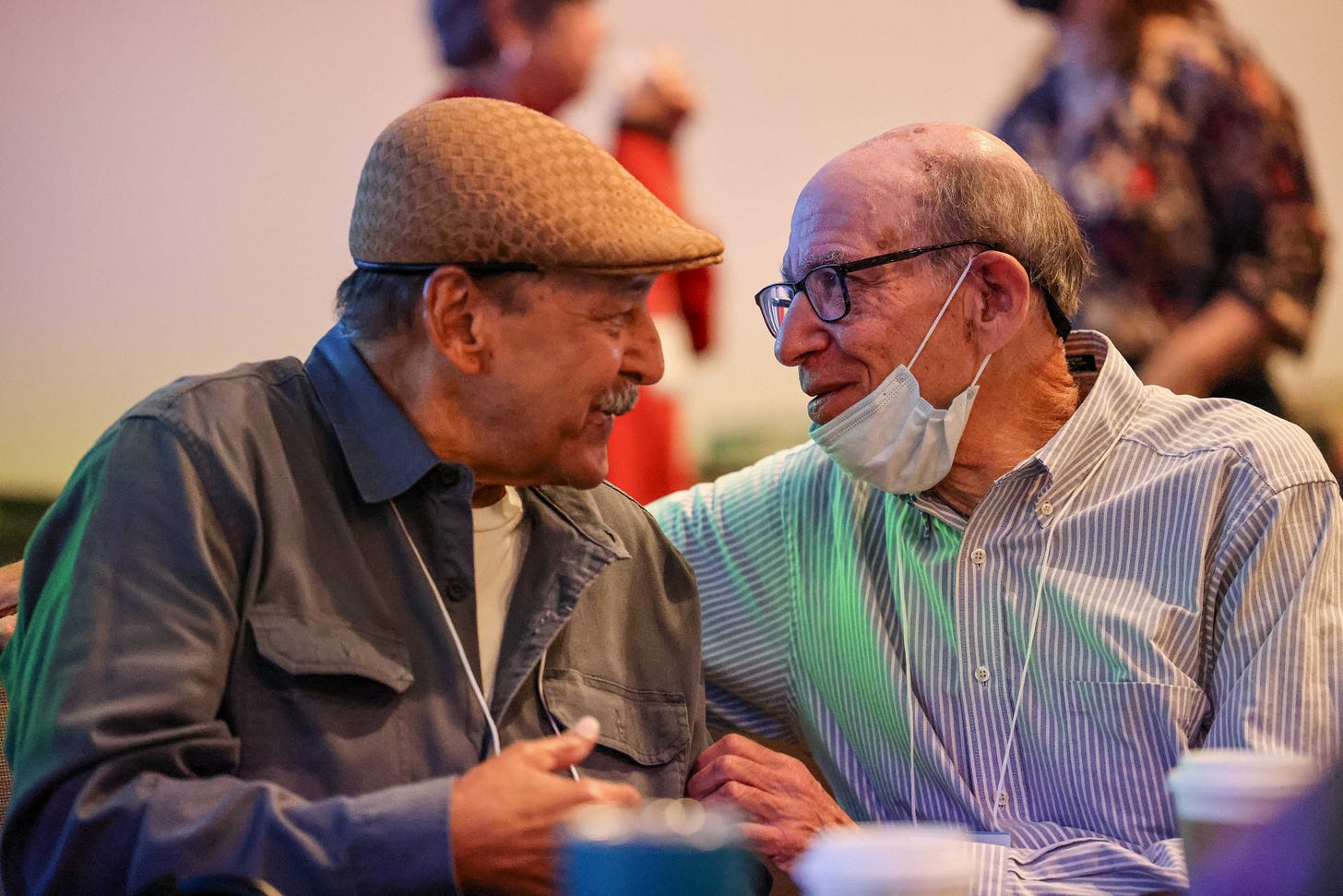 Two men leaning in close, talking to each other, viewed in profile, both older. Max on the right in a collard shirt, Richard on the left in a jaunty hat and with a moustache.