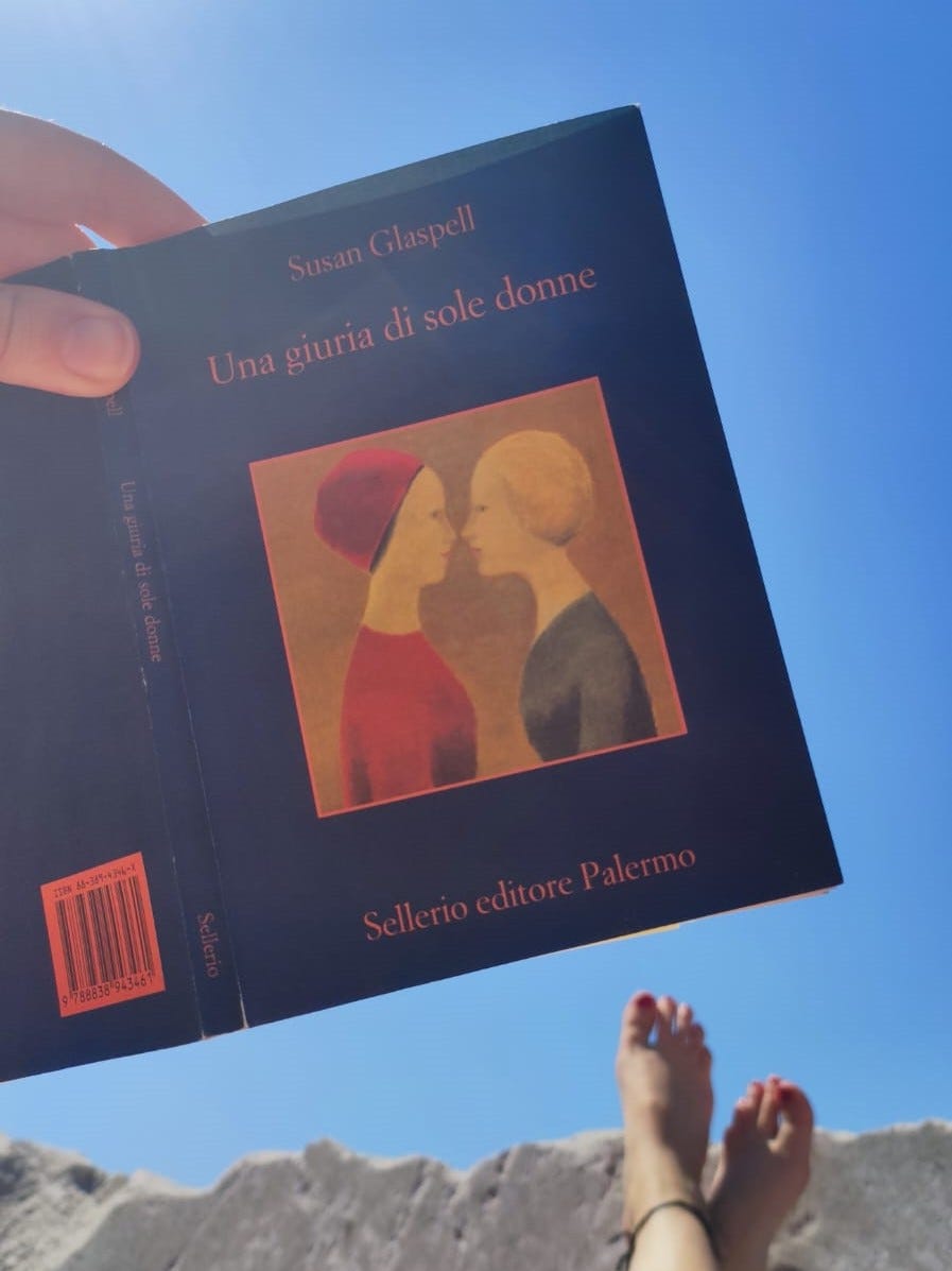 Libro con copertina blu tenuto in mano. Sullo sfondo il cielo azzurro e in basso dei piedi appoggiati a degli scogli