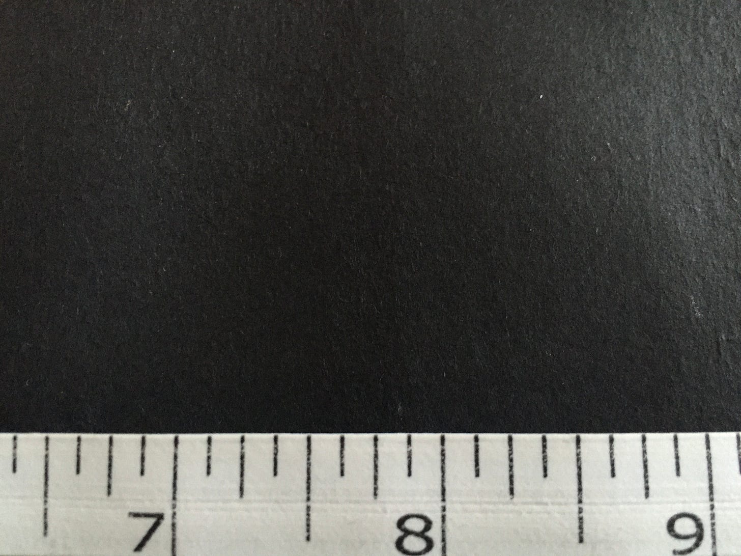A white tape measure with black tick marks and numbers  7,  8 and 9 sits  on a shiny and bumpy black surface.