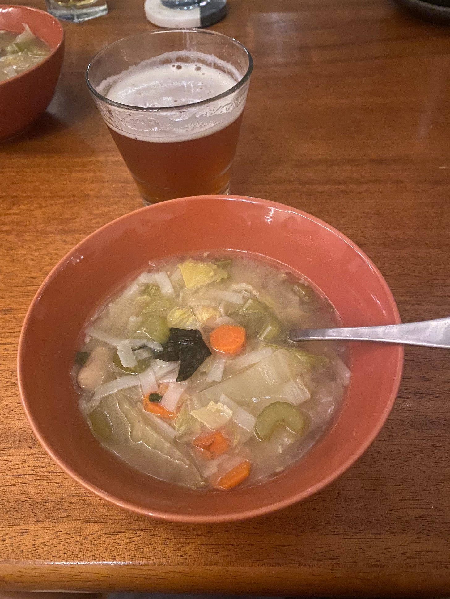 An orange bowl of vegetable soup with flat rice noodles. Behind it is a glass of beer with a little foam on top.