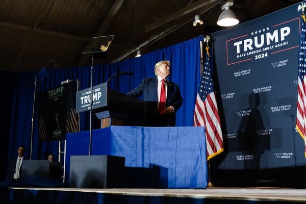 Former President Donald J. Trump stands behind a lectern and in front of a wall bearing Trump 2024 signage at a campaign event in New Hampshire.