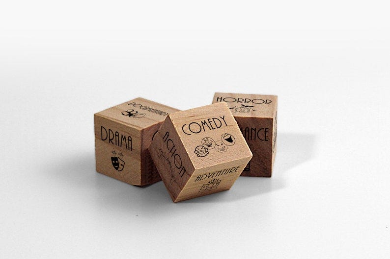 May include: Three wooden dice with different movie genres printed on each side. The dice are brown and have black text. The genres are 'Drama', 'Action', 'Comedy', 'Adventure', 'Horror', and 'Romance'.