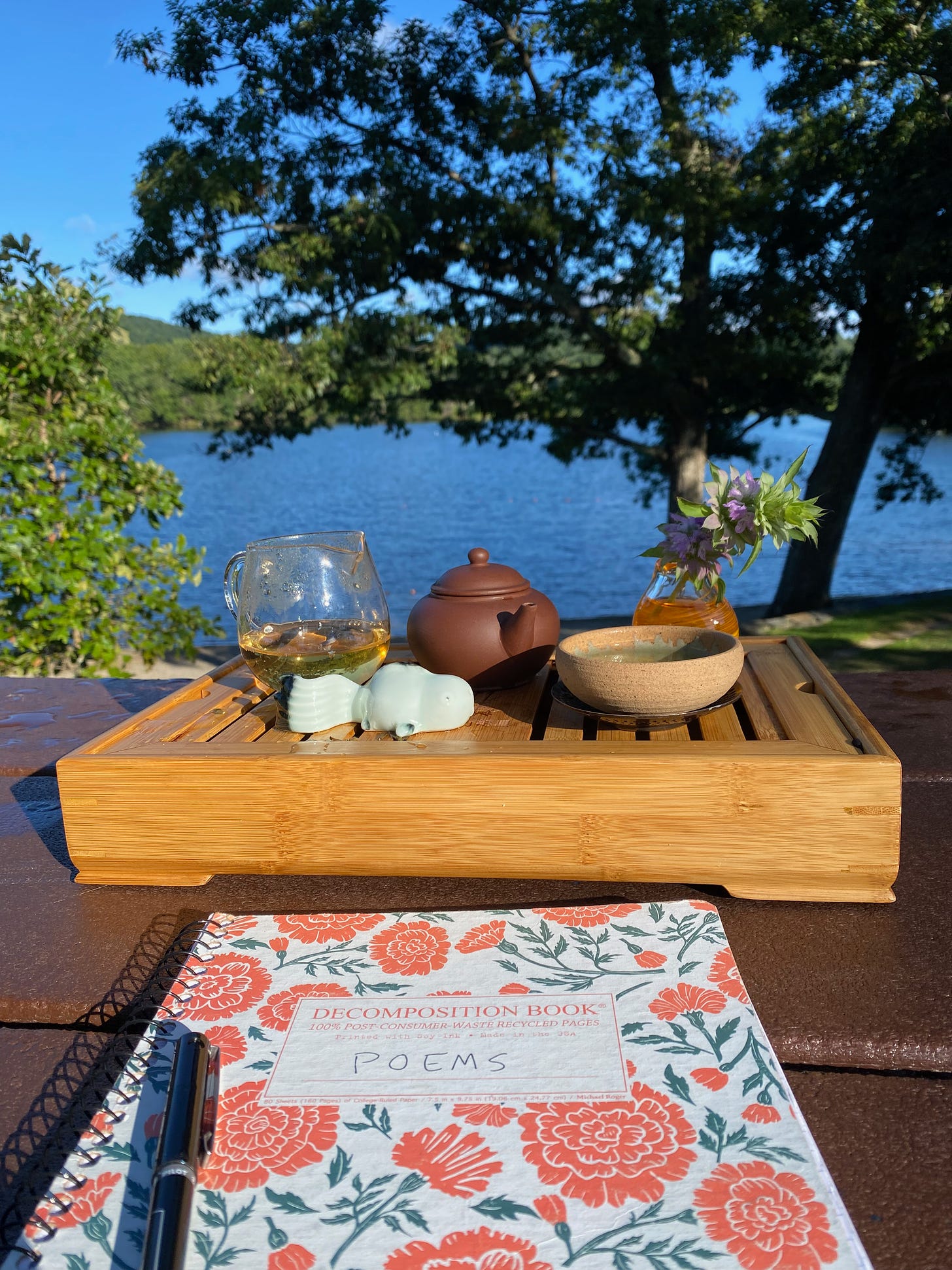 A tea setup on a picnic table overlooking Ashfield Lake. The table also holds my poetry notebook. the water is deep blue, the light bright and sharp. There’s a large oak tree on the shore.