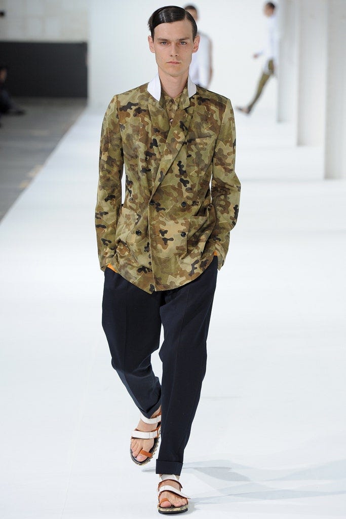 A model on a fashion catwalk, who is a white man with slick dark brown hair, wearing an all-over green camouflage print double breasted jacket with the collars popped up, with navy trousers and sandals with white and orange straps and a chunky sole. A model on a fashion catwalk, who is a white man with slick dark brown hair, wearing an all-over green camouflage print double breasted jacket with the collars popped up, with navy trousers and sandals with white and orange straps and a chunky sole.