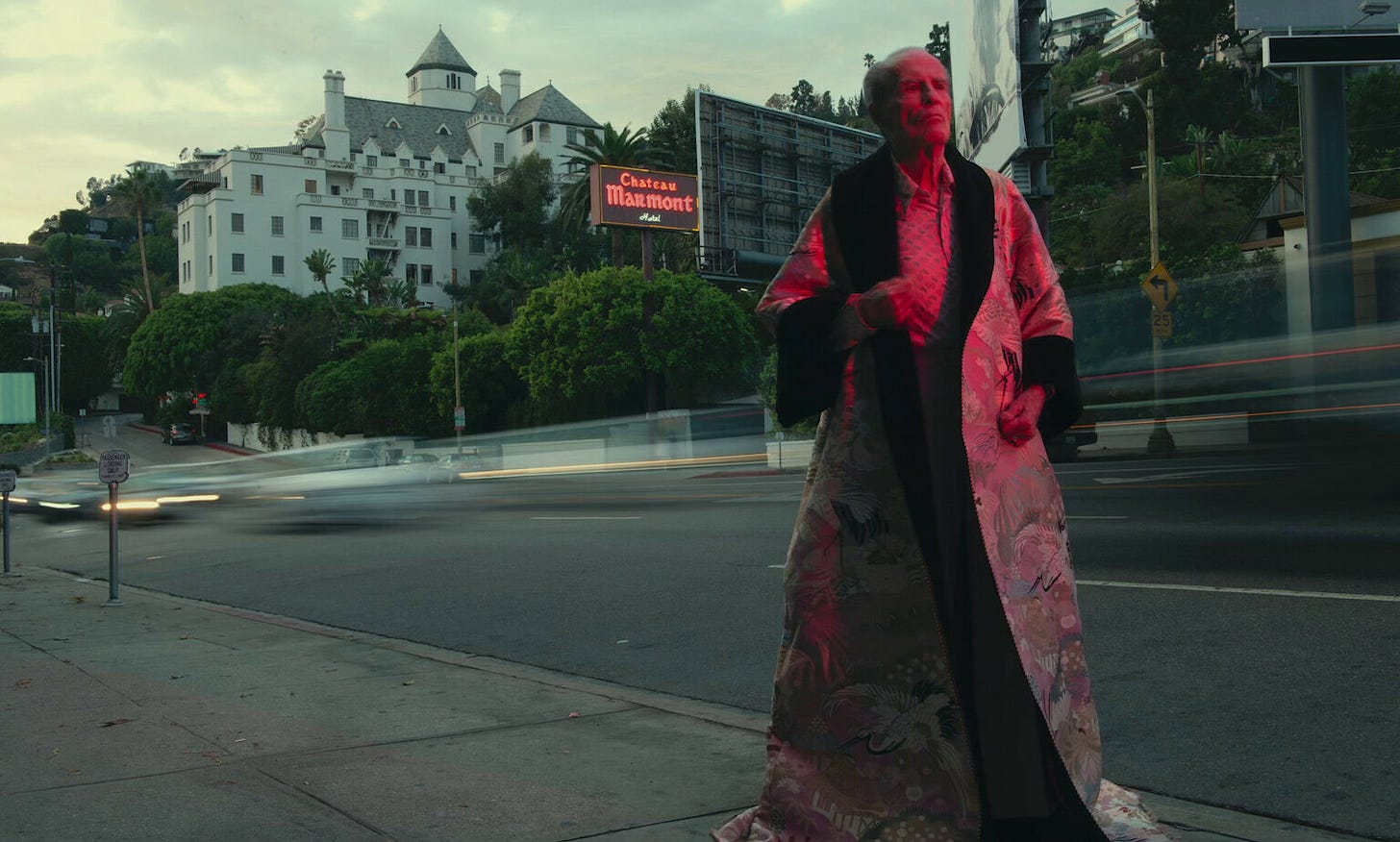 Photograph of film-maker and writer Kenneth Anger, wearing an elaborate brocade cape in the street outside the famous Hollywood Hotel Chateau Marmont. Wikimedia Commons.