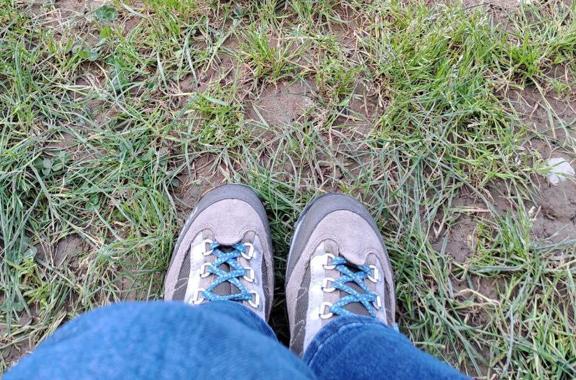 Primo piano di un paio di scarponcini da trekking grigi con lacci blu, indossati da una persona con jeans. I piedi sono posizionati su un terreno erboso con alcune zone di terra visibili.