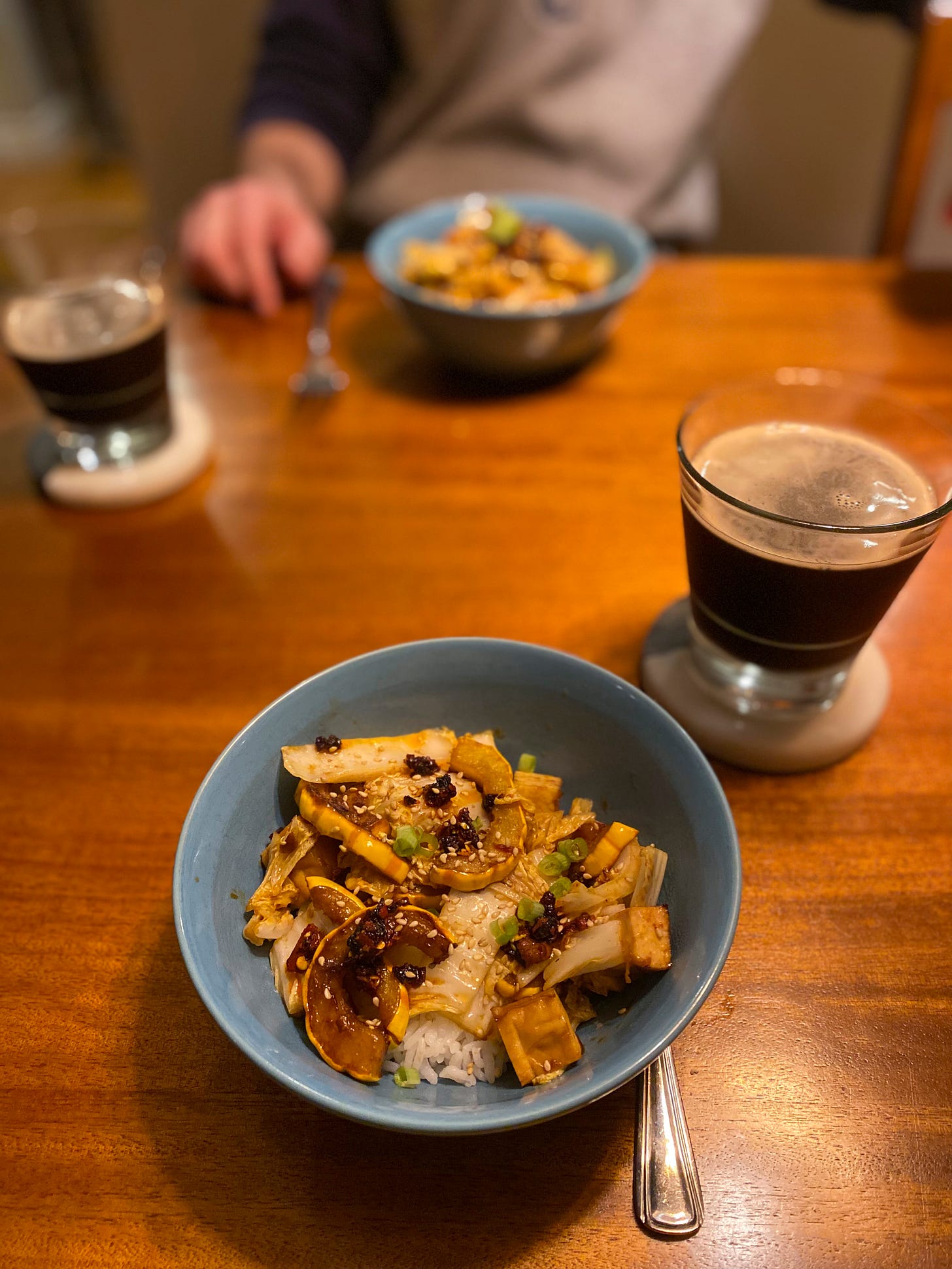 Two blue bowls of rice with braised squash slices, pieces of napa cabbage, and cubes of tofu in hoisin glaze. On top is chili crisp and sesame seeds, and two glasses of dark beer rest on coasters beside the bowls.