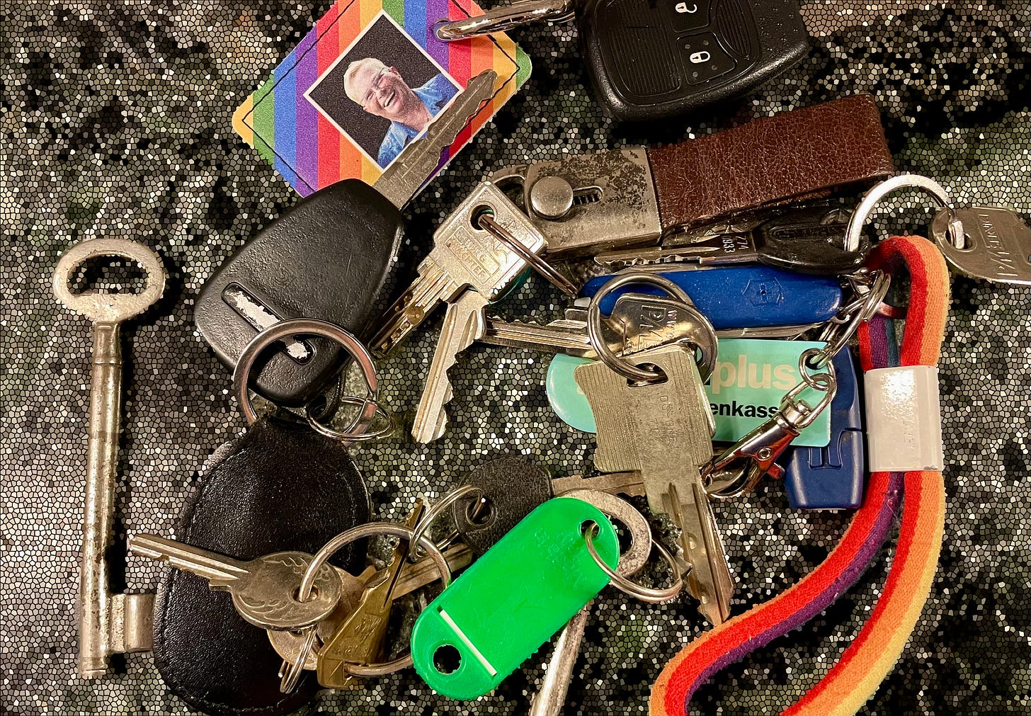 A set of keys on a mosaic-textured surface, including car keys, a Jeep key fob, a rainbow keychain with a photo, a Cape Point-South Africa medallion, and various house keys.