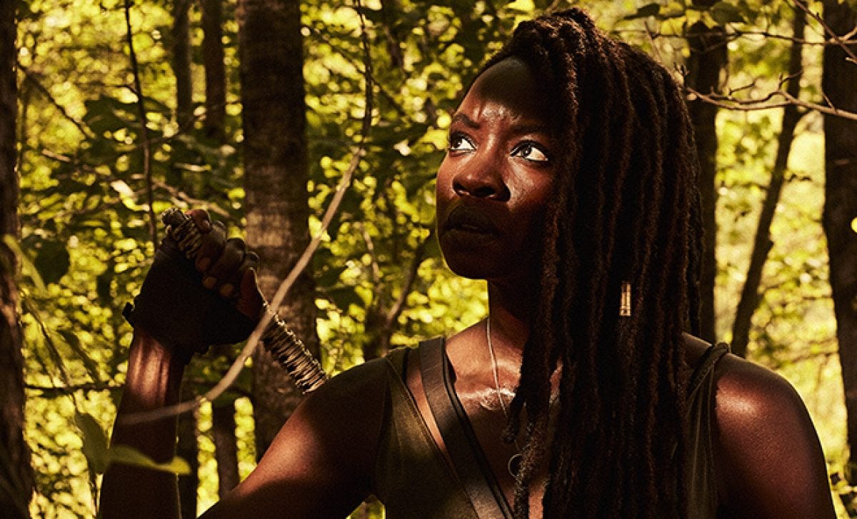 Michonne (Danai Gurira): numa floresta, mulher negra segura a base da espada presa em suas costas