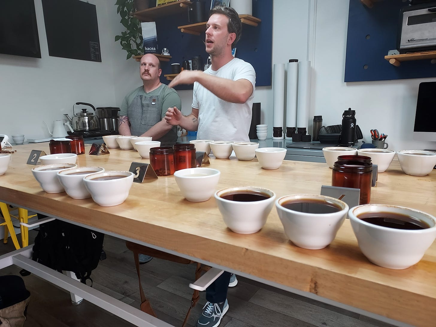 A teacher standing behind a table filled with coffee cupping bowls gestures wildly while talking about coffee. A mustachioed gentleman looks on.