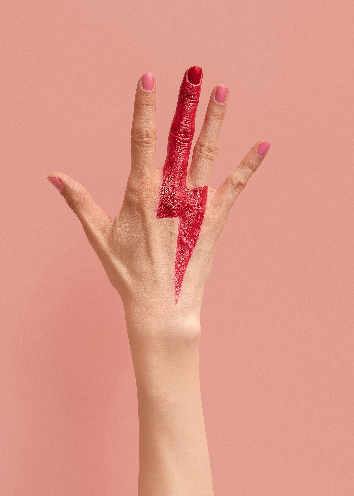 A woman's hand raised against a pink background. The middle finger is painted with a David Bowie-style red lightning bolt. 