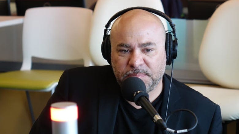 A bald man with a short grey beard sits in front of a microphone wearing a dark blazer over a black shirt, with black headphones on his head.