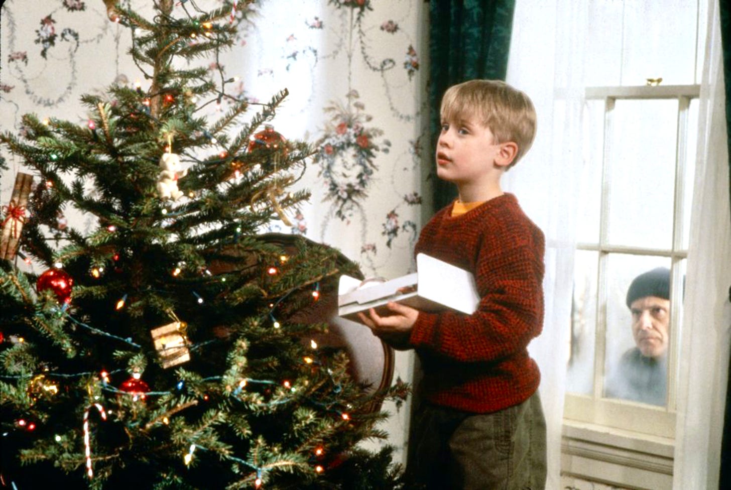 A burglar peeks through a window at a young boy decorating a Christmas tree.