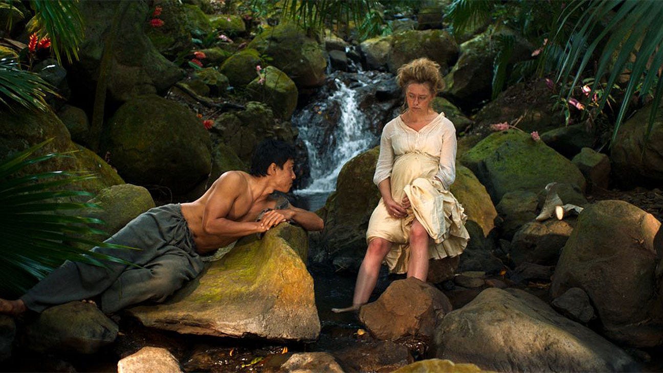 A shot of a topless Indonesian man Reza (Muhammed Khan) reclining on a rock next to a waterfall and gazing at Josefien (Lisa Zweerman), a Dutch woman wearing a white dress