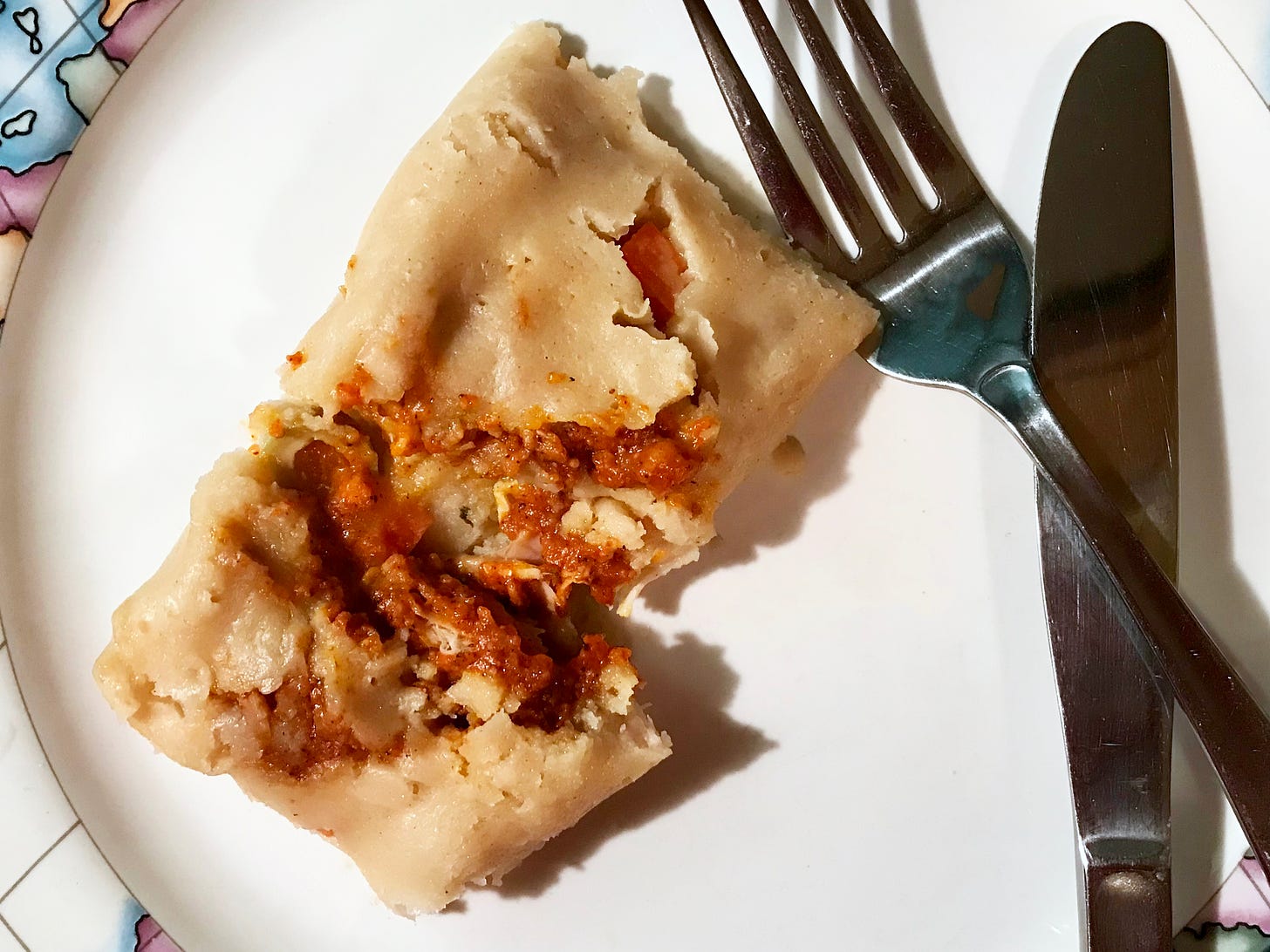 An unwrapped tamale on a plate with a knife and fork. The tamale is cut so the filling cane be seen.