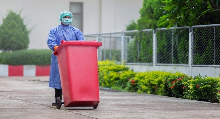 a healthcare worker in full PPE wheels a red garbage can down a paved street
