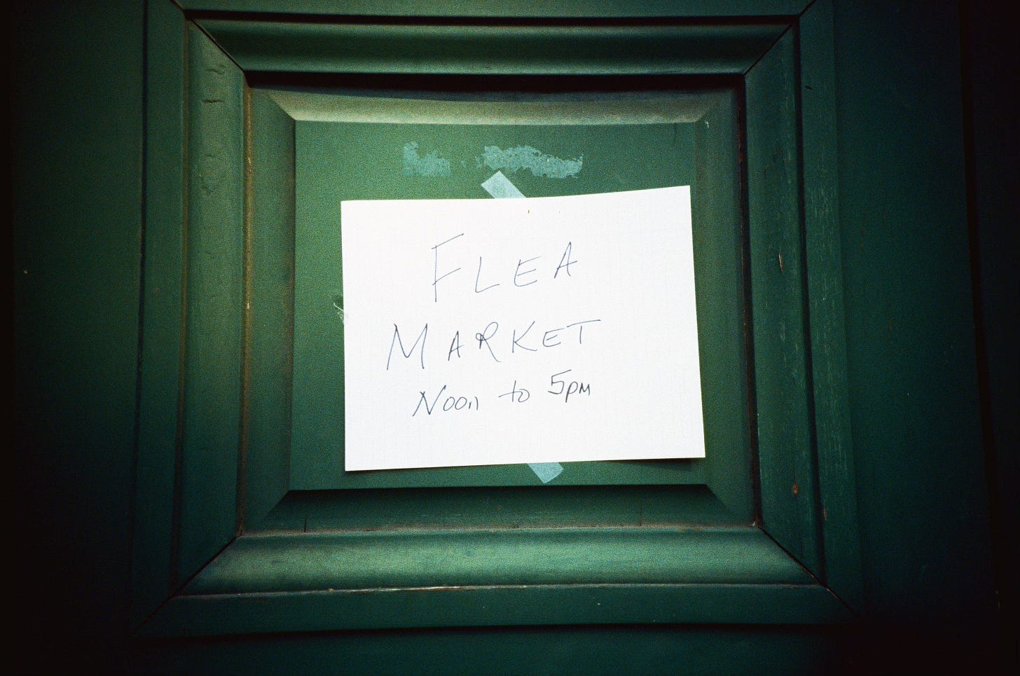 a paper sign on a green door reading, "Flea Market Noon to 5pm"