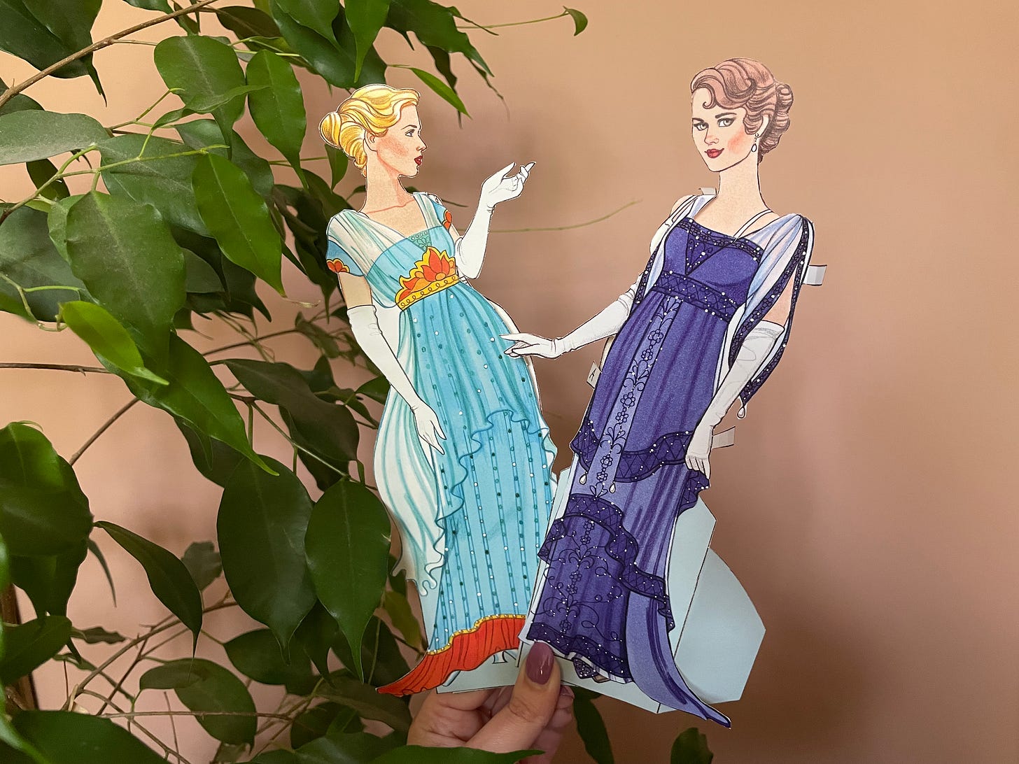 Two paper dolls in Belle Epoque style, beaded evening gowns, held in front of a green leafy plant and a terracotta pink wall.