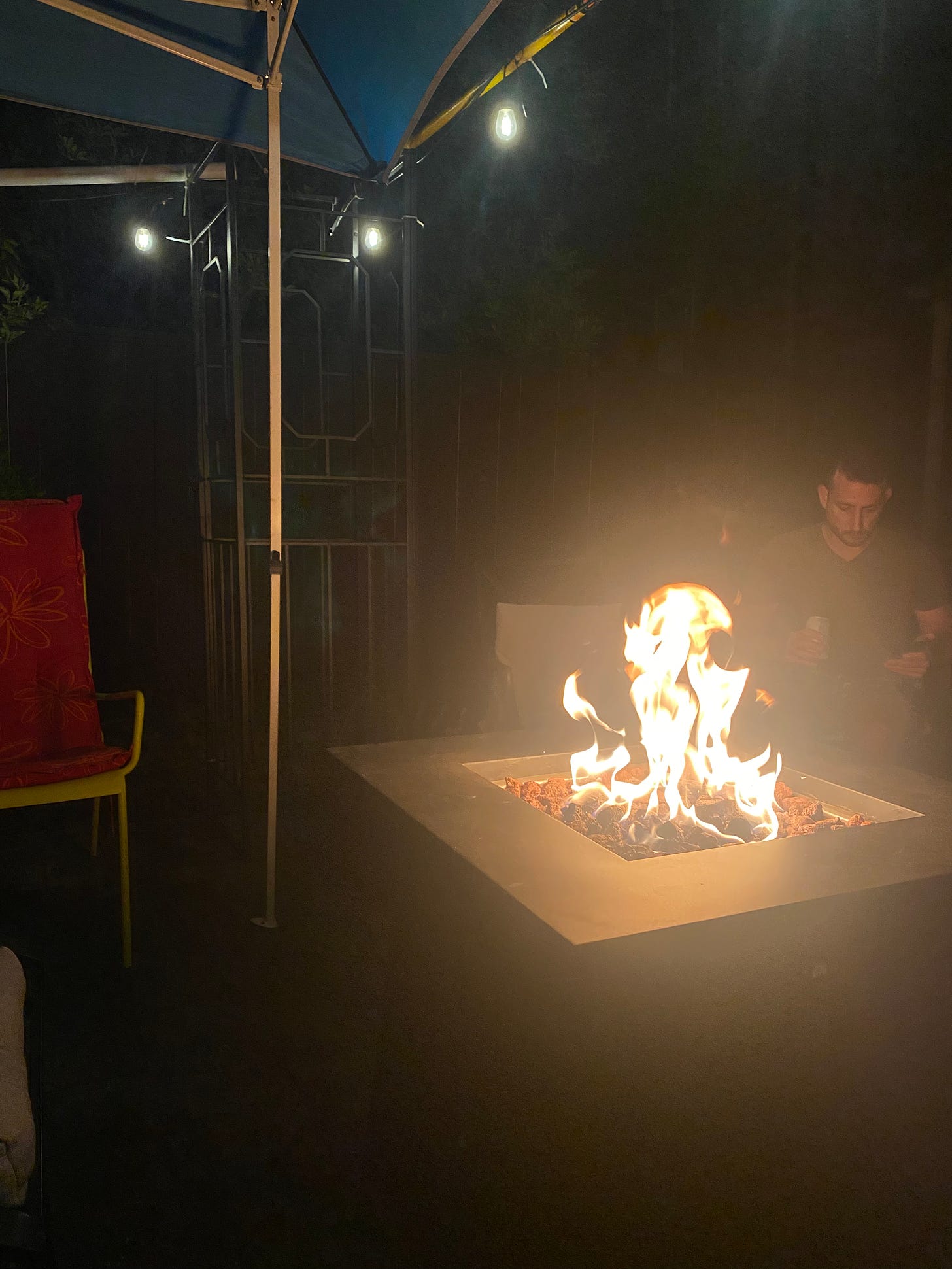 A backyard at night, patio chairs around a fire table.