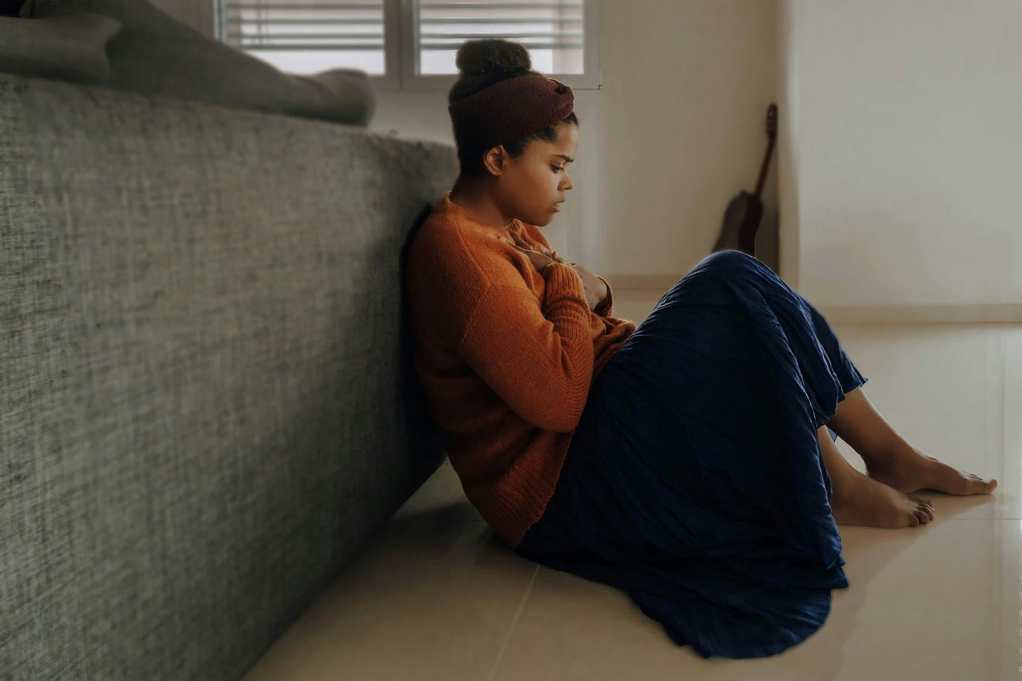 Young Black woman sitting on the ground in a bedroom with her back against the bed and clutching her hands to her chest in grief