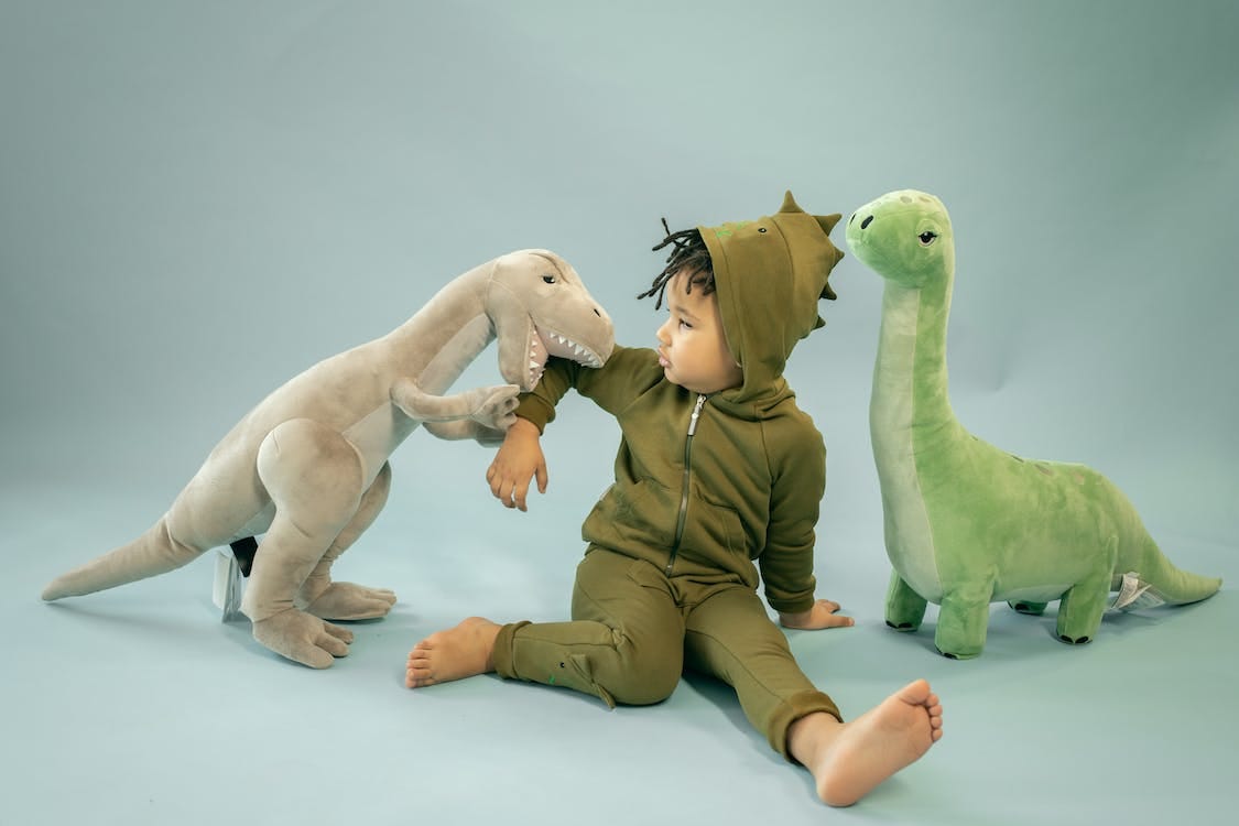 Free African American child with dreadlocks in dinosaur costume sitting between soft toys representing bite concept Stock Photo