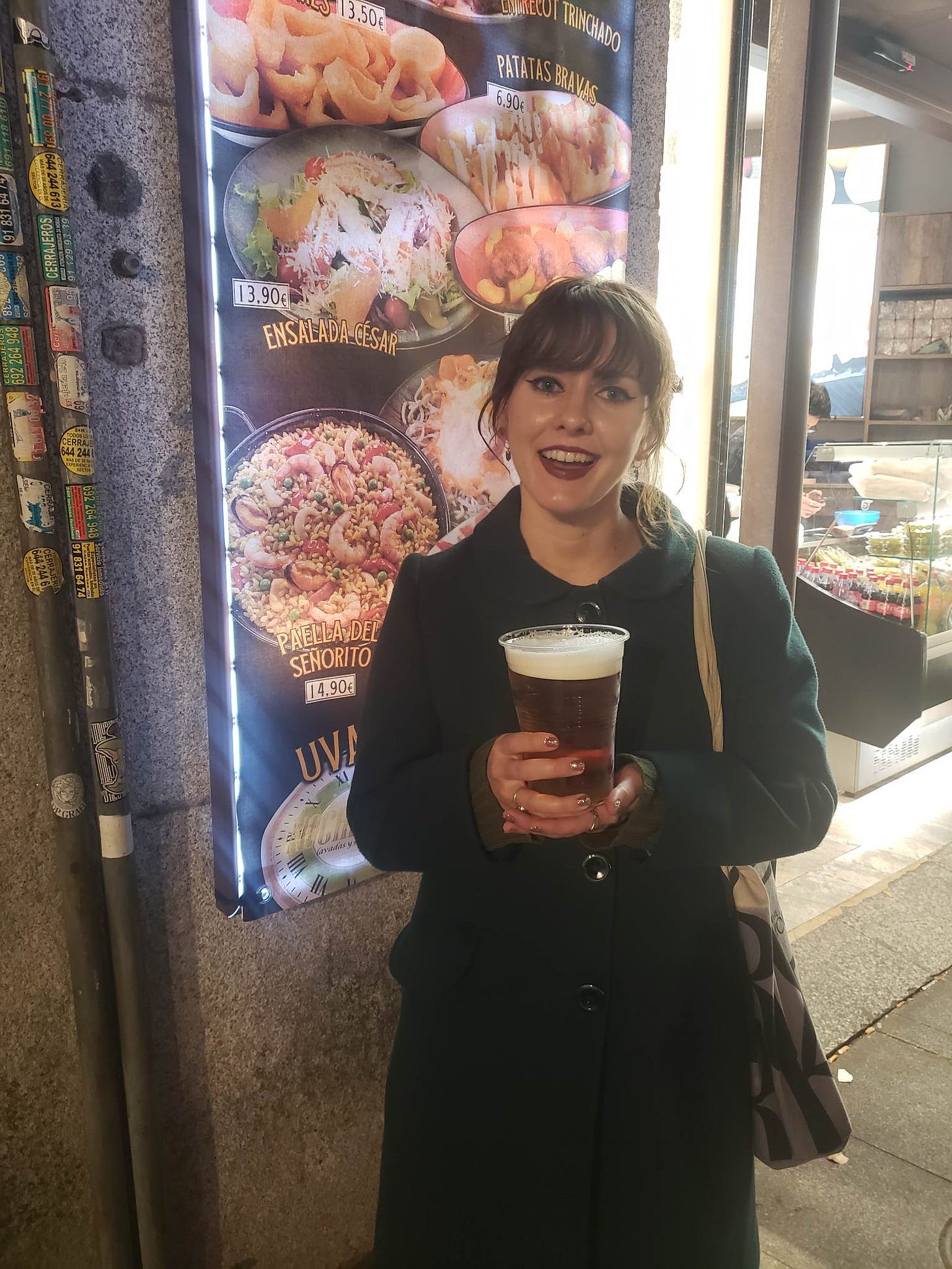 A young woman smiles and stands outside a restaurant. She is holding an enormous cup of beer, maybe three times the size of a normal beer on tap.