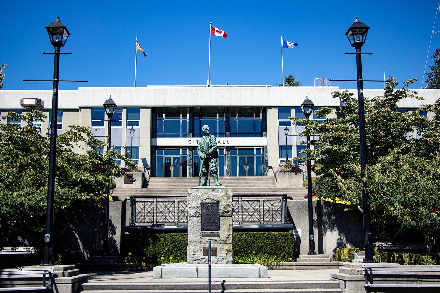 New Westminster City Hall is in the background, with the words "city hall" obscured somewhat by a statue in the foreground, about a storey below. the photo is taken from a plaza where the statue stands, which is lush with trees and other plant life. atop city hall are three flags: canada's, BC's and i think the city's?