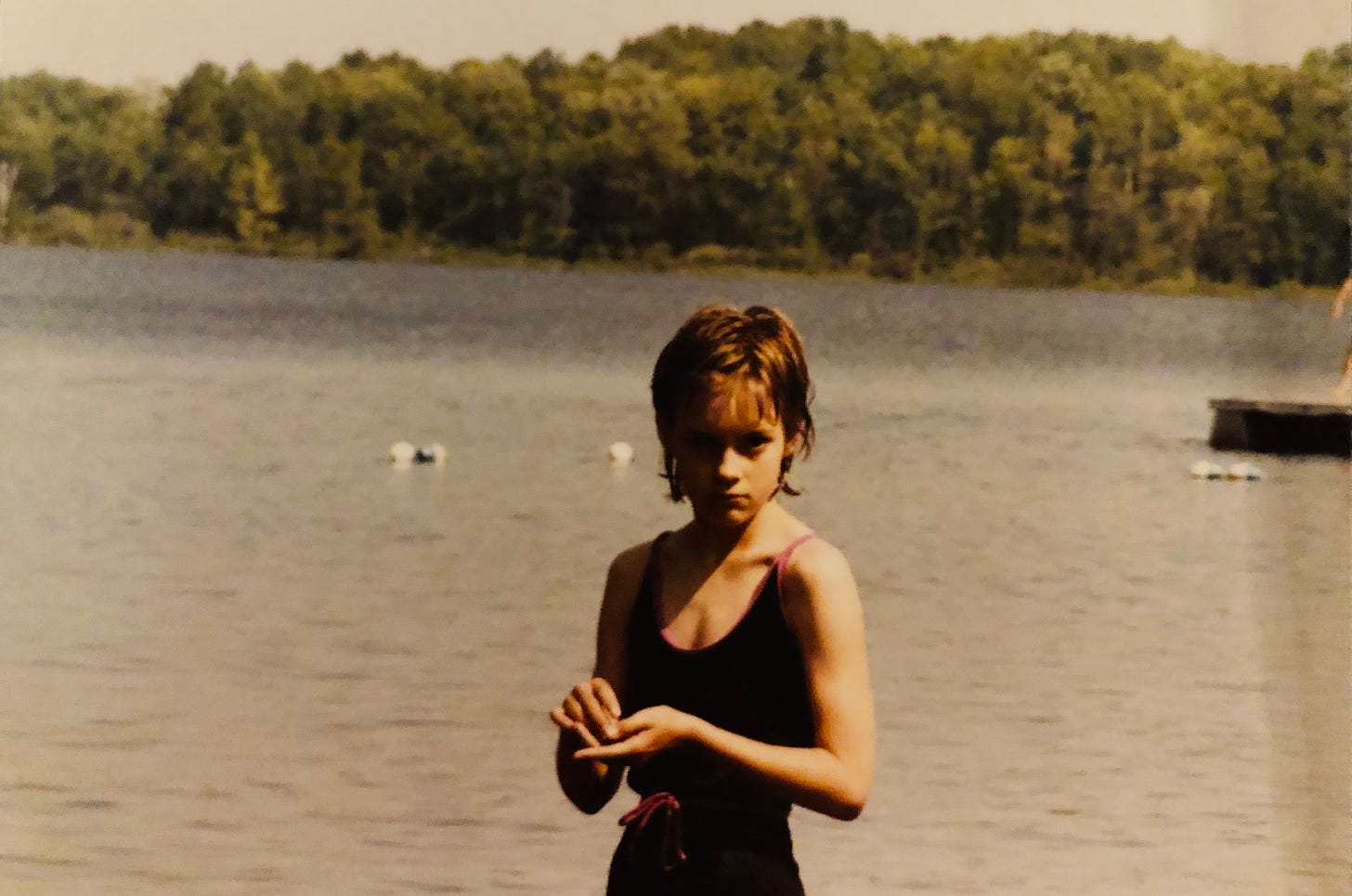 A grouchy twelve-year-old girl in a black swimsuit glares at you from the shore of a lake with swimming buoys and trees in the background