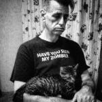 Johnny in burnout. Personal photo. Johnny Profane, early 60s, in a black t-shirt that reads, “Have you seen my zombie?” He holds a cat.