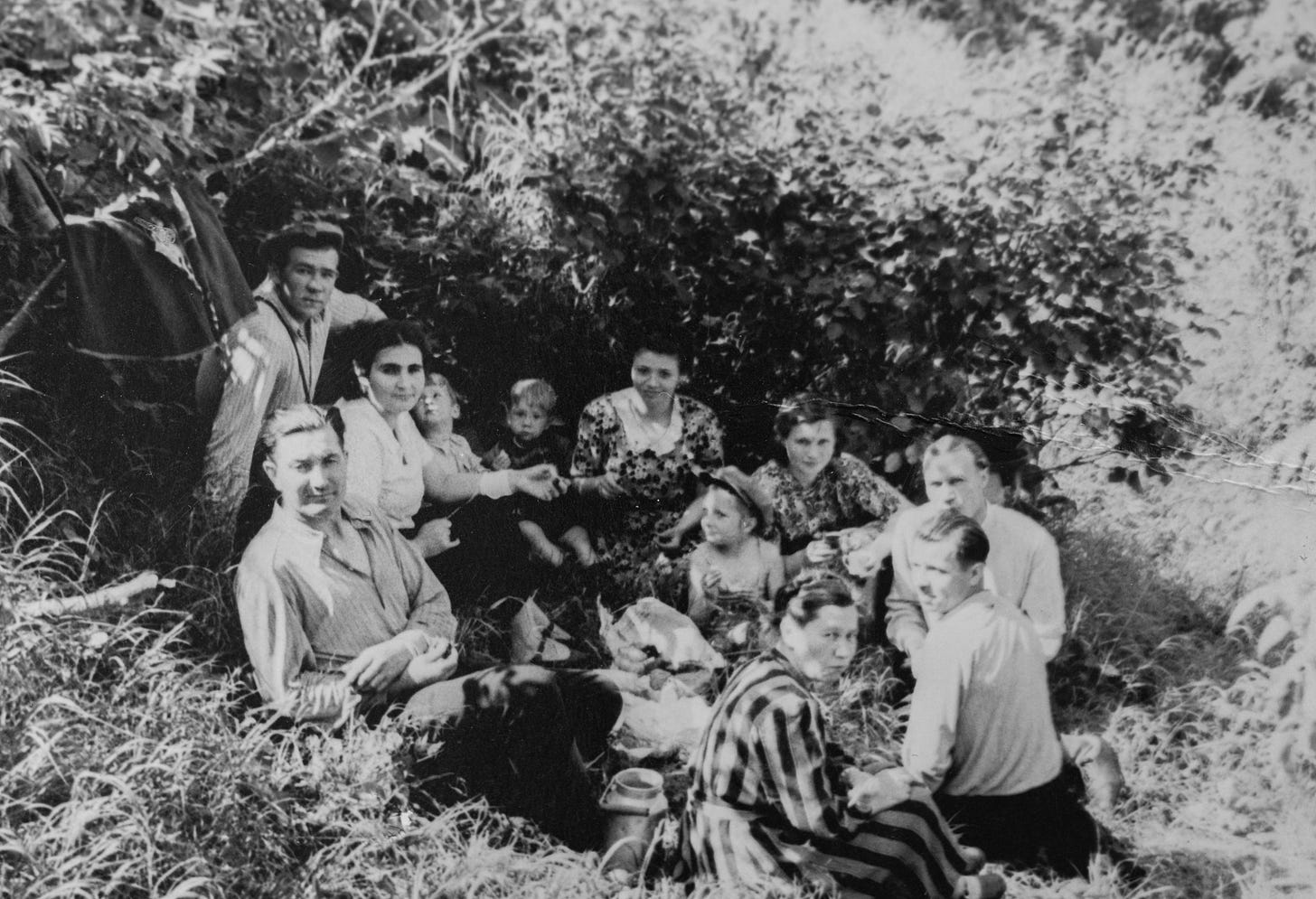 A black and white photograph of a family gathering in a rural setting, men, women and children sitting on the grass, smiling to the camera, maybe they gathered for a picnic. They seem relaxed, having a good time together.