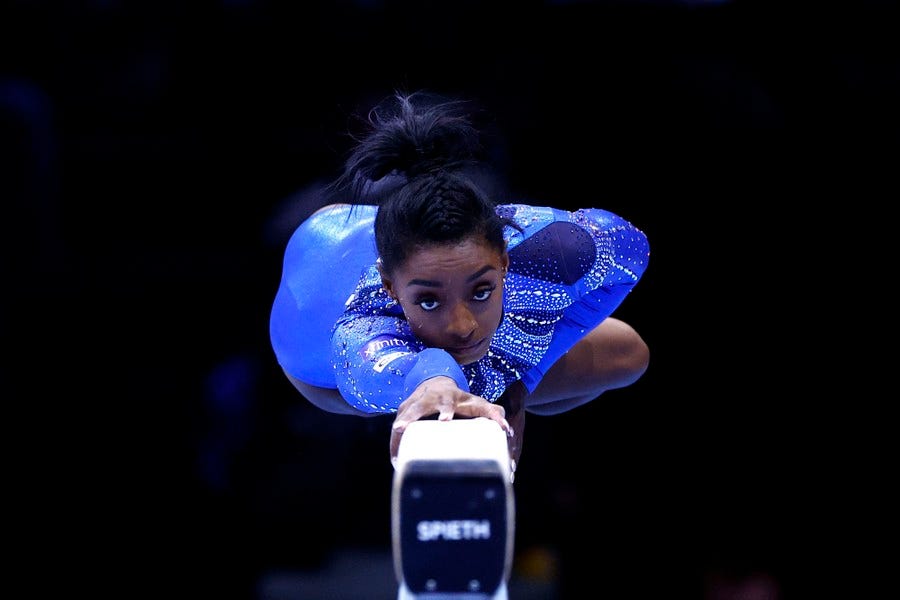 A gymnast lies stretched-out atop a balance beam wearing a shiny blue leotard