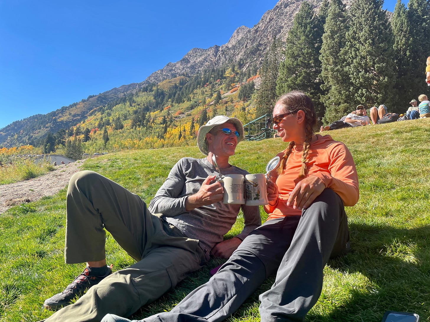 Grice and Mina sitting on a grassy slope with mountains in the background, holding beer steins in hand and cheering.