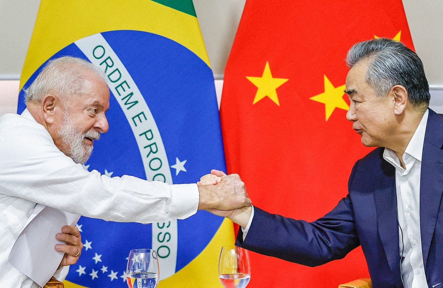 Imagem do presidente Lula vestindo camisa branca e apertando a mão do chanceler chinês Wang Yi, com terno azul marinho e camisa branca, sem gravata. Ao fundo estão bandeiras brasileira e chinesa. Copos de água abaixo deles