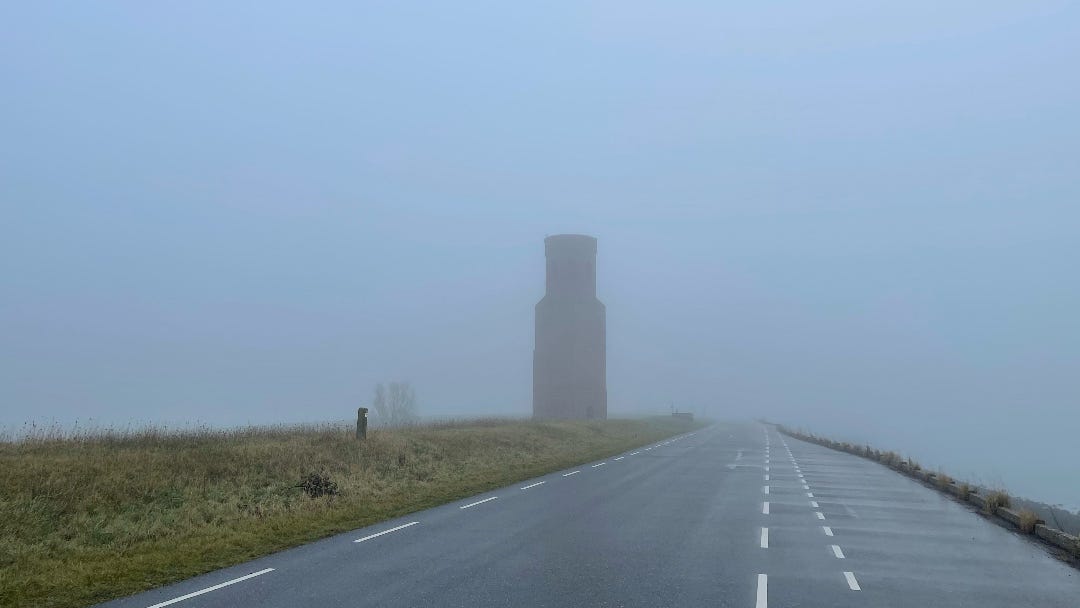 photo as described in the text: misty, gray, a road disappearing into the mist with on the left side of the road still visible a lonely church tower.