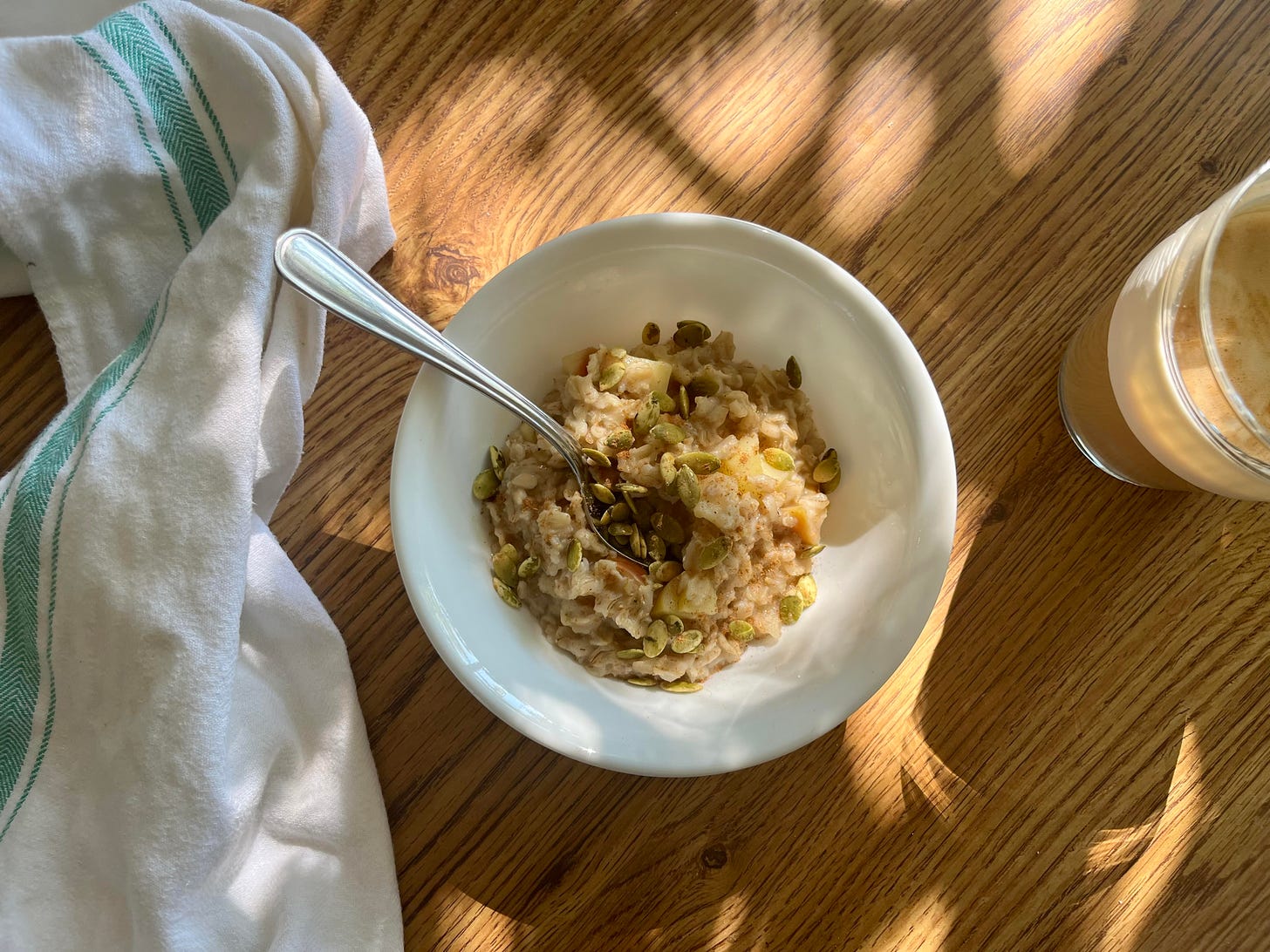 A flat lay image of a bow of oatmeal sprinkled with pumpkin seeds, a glass of coffee and a white dish towel