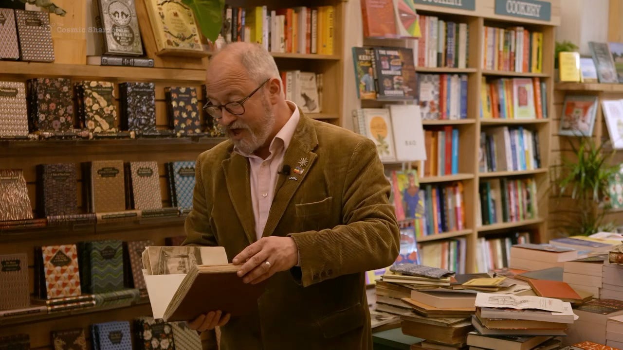 Robin Ince holding an old scrapbook in a bookshop, surrounded by books