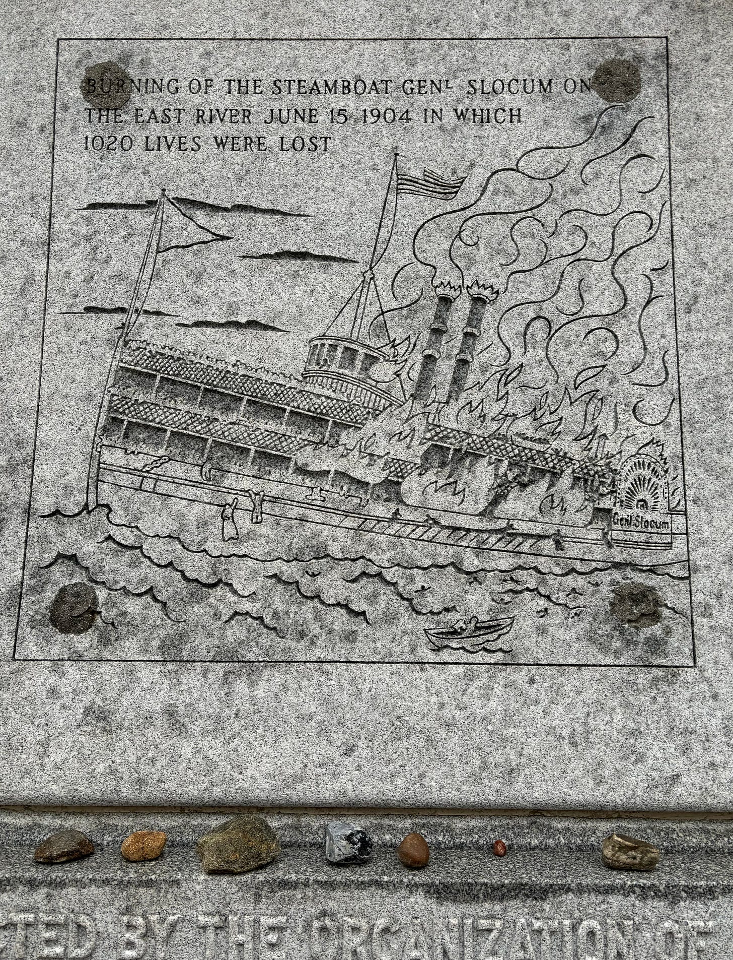 A carved illustration of a paddle ship burning as the stern sinks into the water. It is engraved: "Burning of the Steamboat General Slocum On The East River June 15 1904 In Which 1020 Lives Were Lost. In the illustration, passenger jump from the flames and cling to the rails.In the ledge beneath it sit stones left by those remembering their dead.