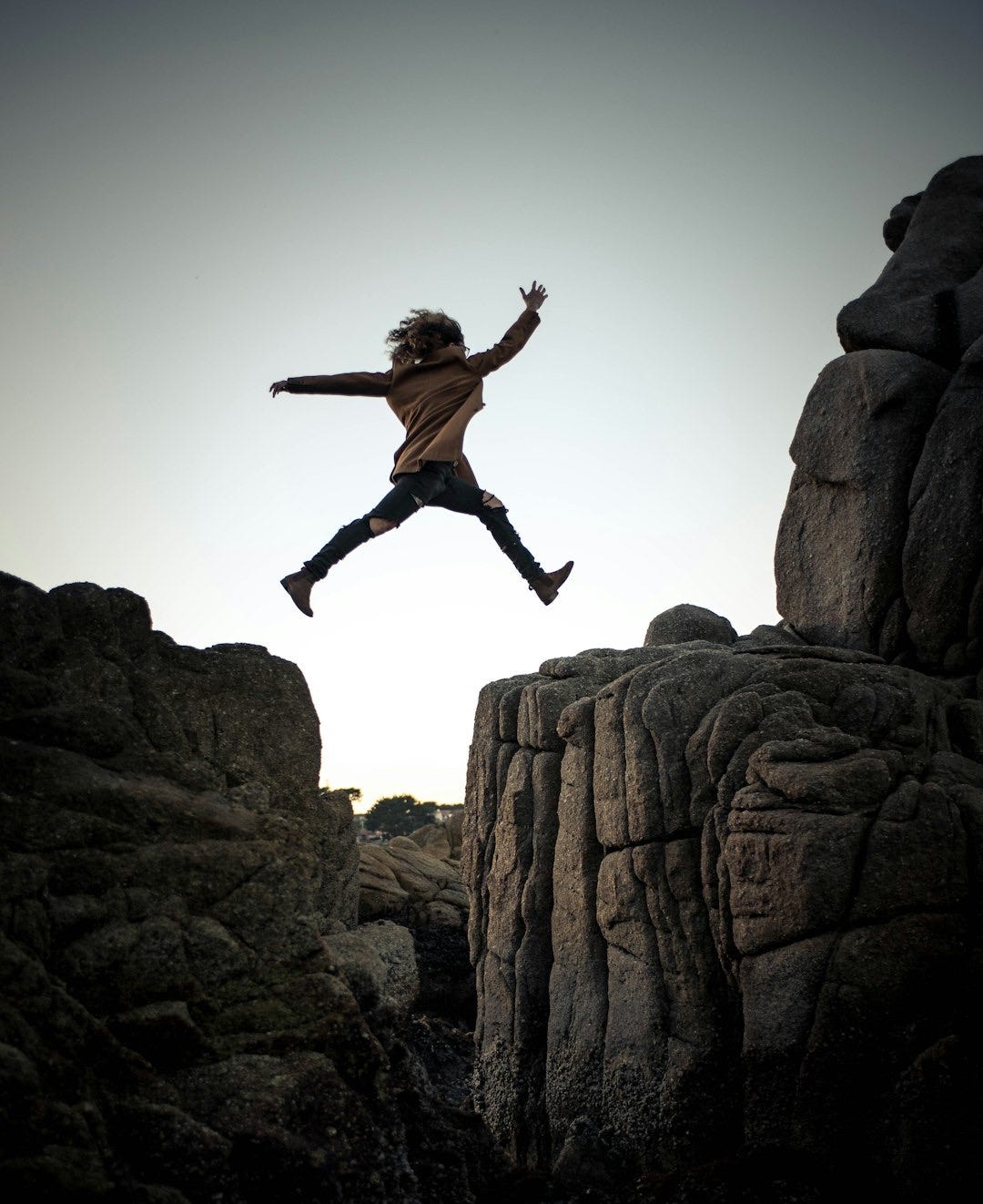 person jumping on big rock under gray and white sky during daytime