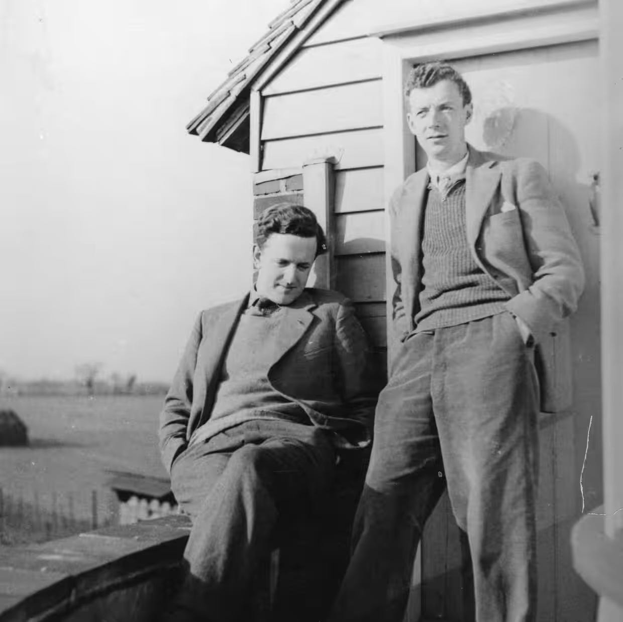 Black and white photo of two men, one seated and one standing, against a small house, with a field and sky visible in the background.