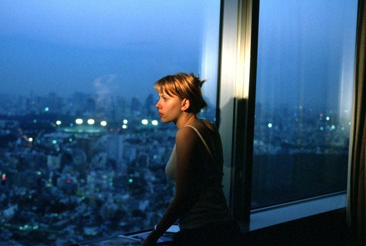 This may contain: a woman standing in front of a window looking out at the city lights and buildings