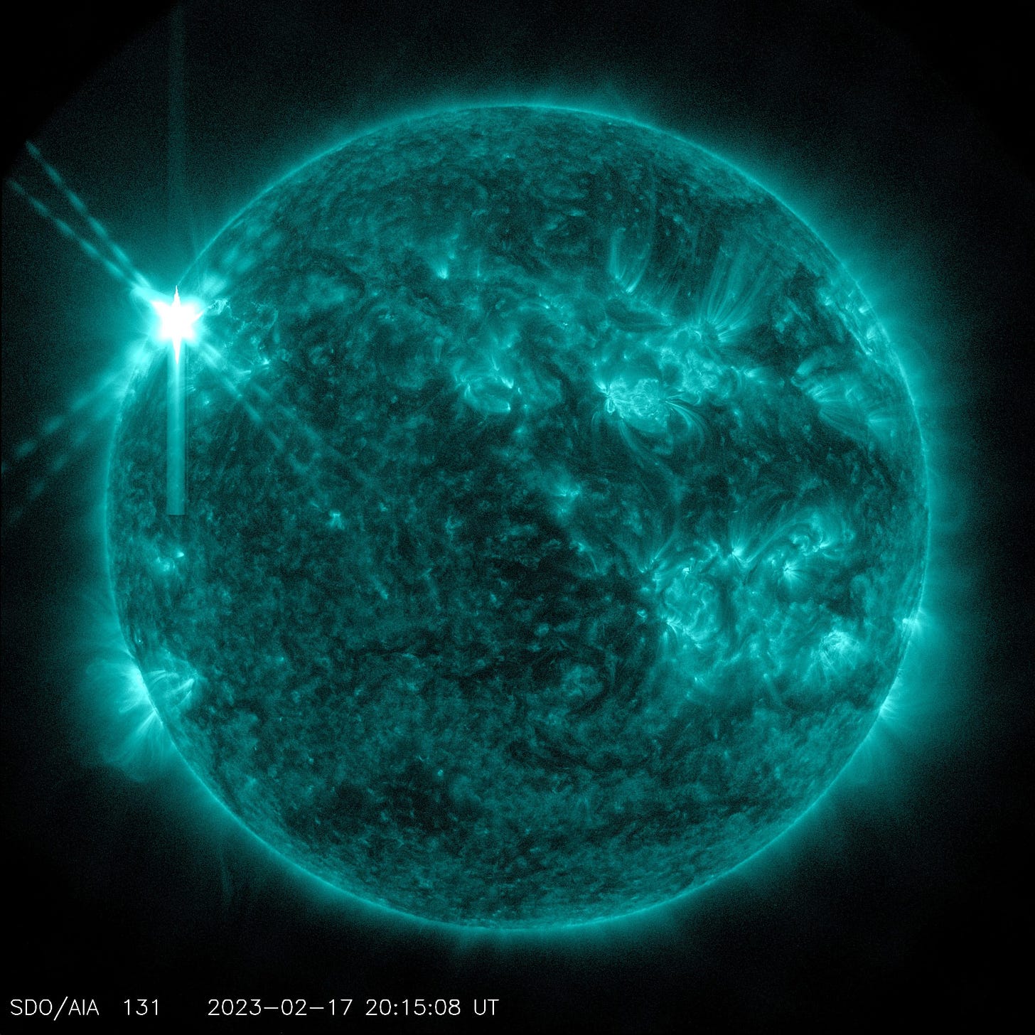 A teal Sun on a black background. The Sun has bright teal areas scattered across the star. On the top left of the star, there is a white X where the flare burst from the Sun.