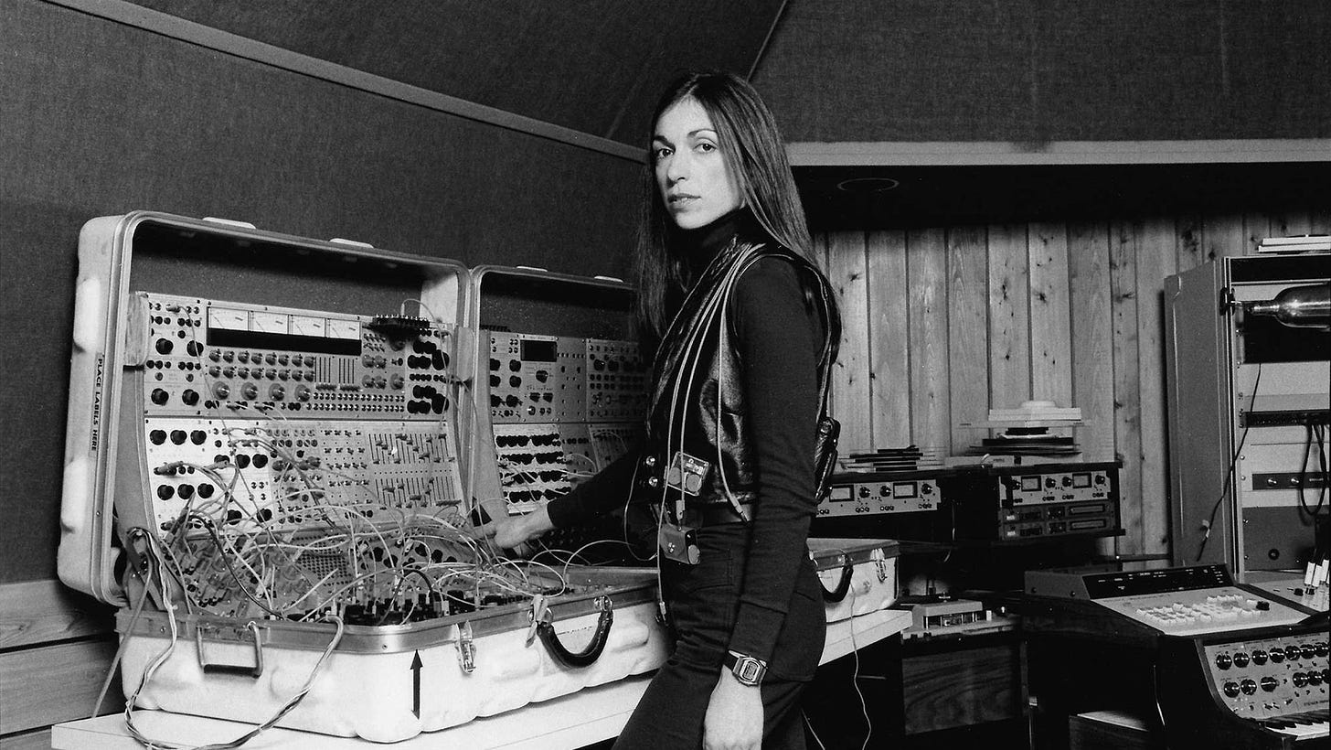 Imagem: Mulher jovem, cabelos pretos, lisos e compridos em frente a diversos sintetizadores modulares com cabos para todos os lados