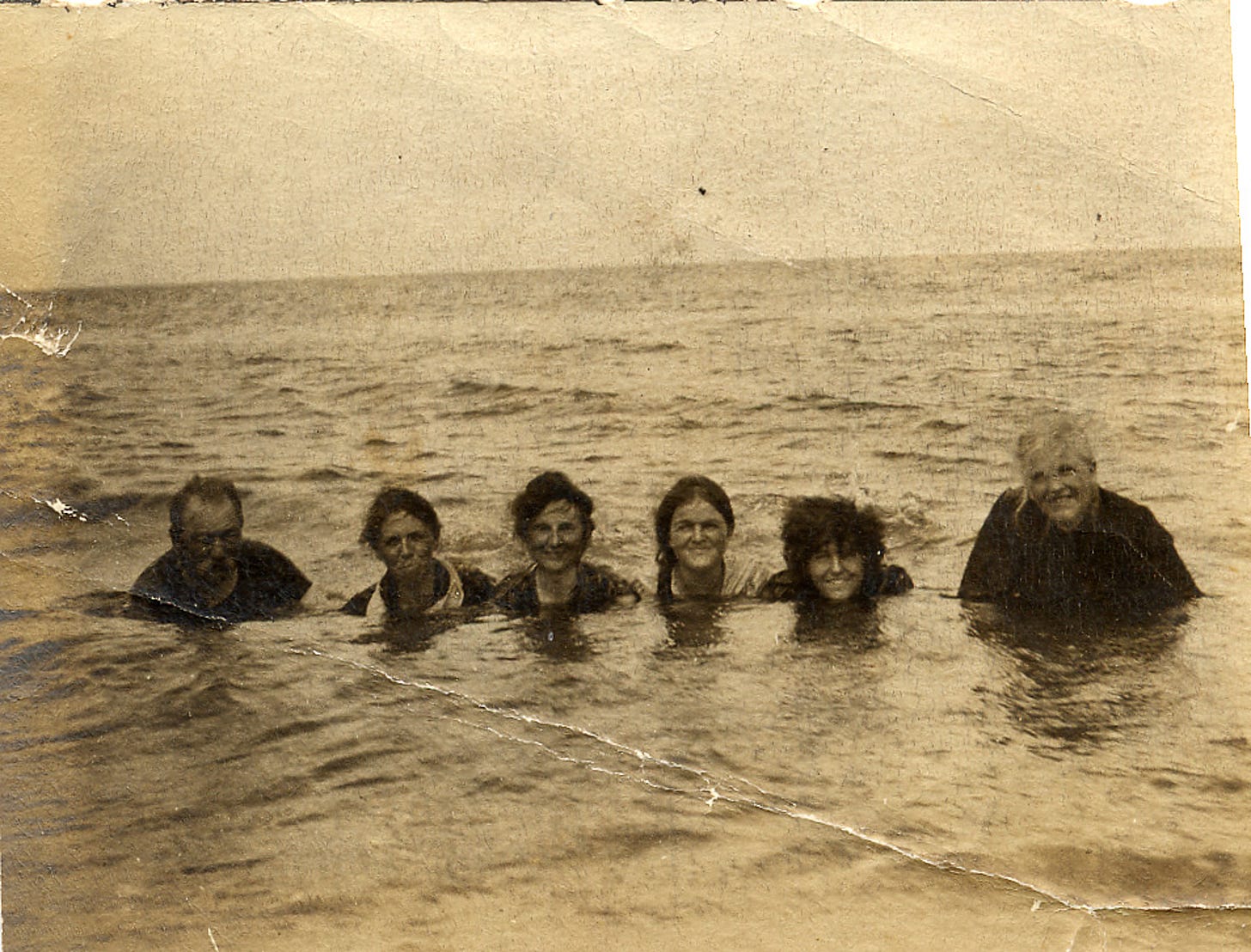 A black and white photo of six people sitting in the water, looking at the camera.