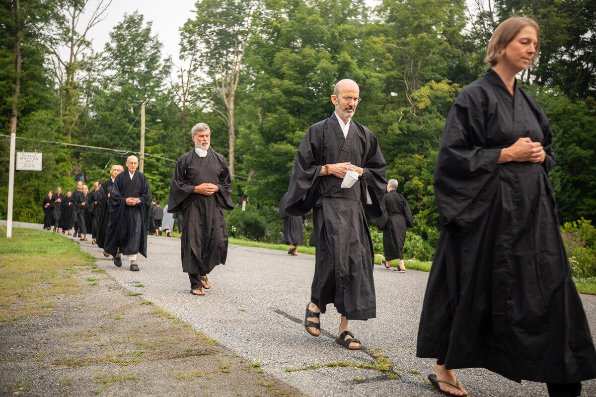  A line of Zen practitioners in black robes practices walking meditation against a bacground of deep green trees.