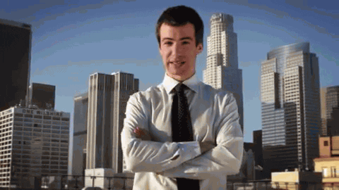 Gif of a white man in business attire (TV comedian Nathan Fielder) smiling, with his arms crossed, in front of a cityscape, as the logo for the TV show Nathan For You flies in from the sides.
