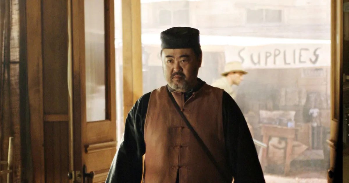 This image shows Mr. Wu (played by Keone Young), the leader of Deadwood's Chinese community, entering through the Gem Saloon's front doors. A sign bearing the word "Supplies" and a white Deadwood resident can be seen in the street behind Wu.