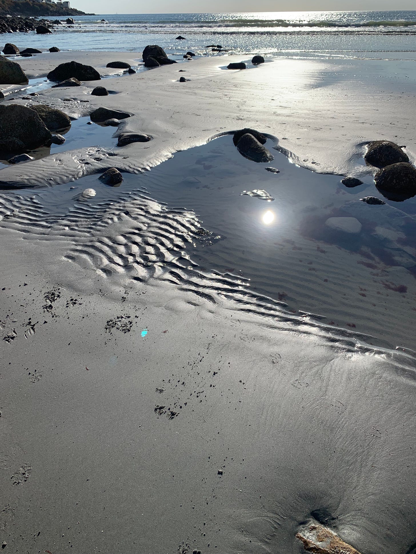 A beach in the morning. The sun is visible reflecting in  tide pool only. The ocean is glimmering in the background, there are rocks around and a rocky coastline to the left.