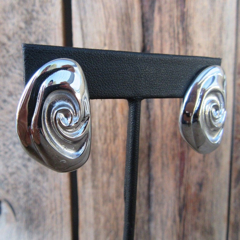 May include: A pair of silver earrings with a swirl design. The earrings are displayed on a black stand.