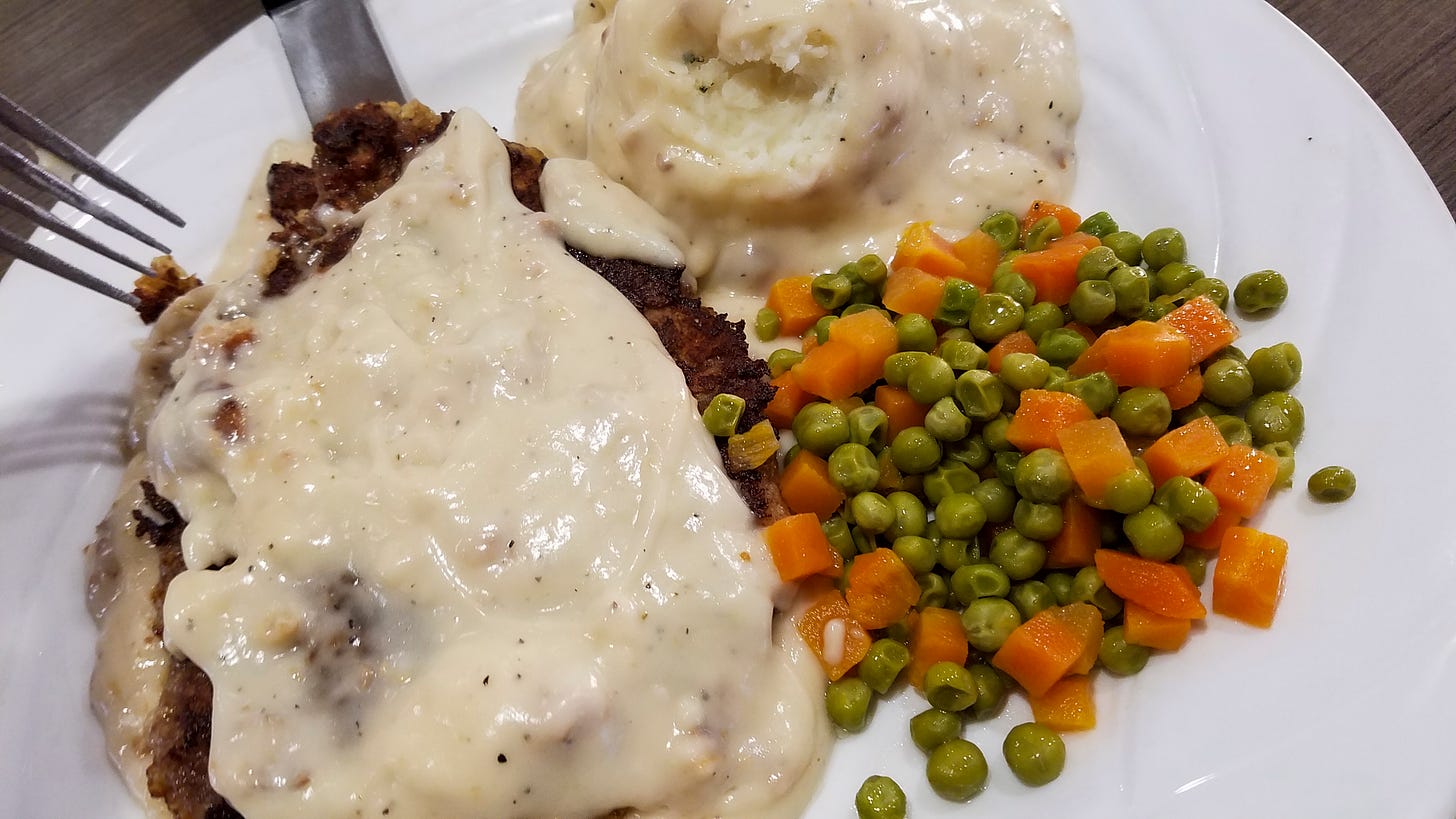 Photo of a plate of food - chicken fried steak with white gravy on top, mashed potatoes with more white gravy, and peas and carrots that look like they came from a can.