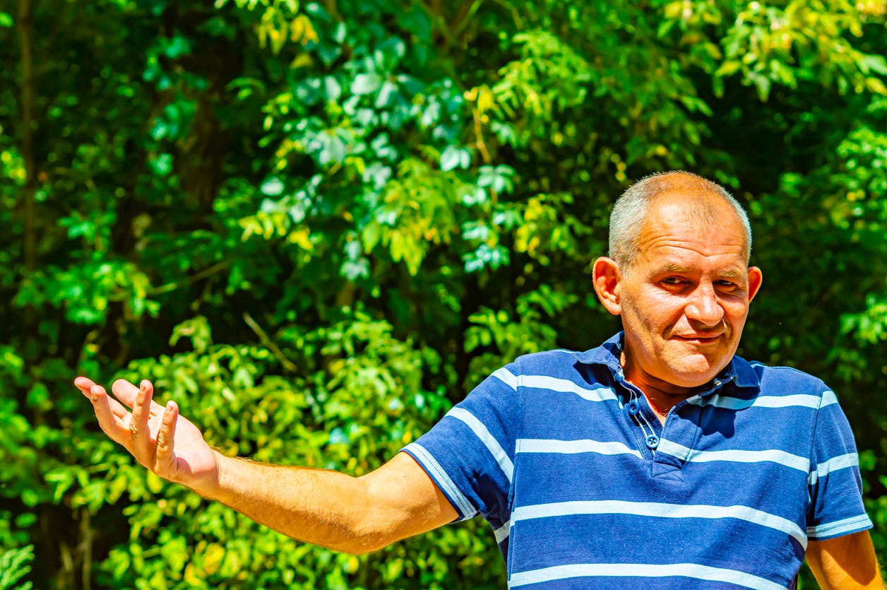 Older white man with gray hair in blue striped shirt against background of greenery in outside sunshine. He's reaching out and motioning with his right arm and hand.